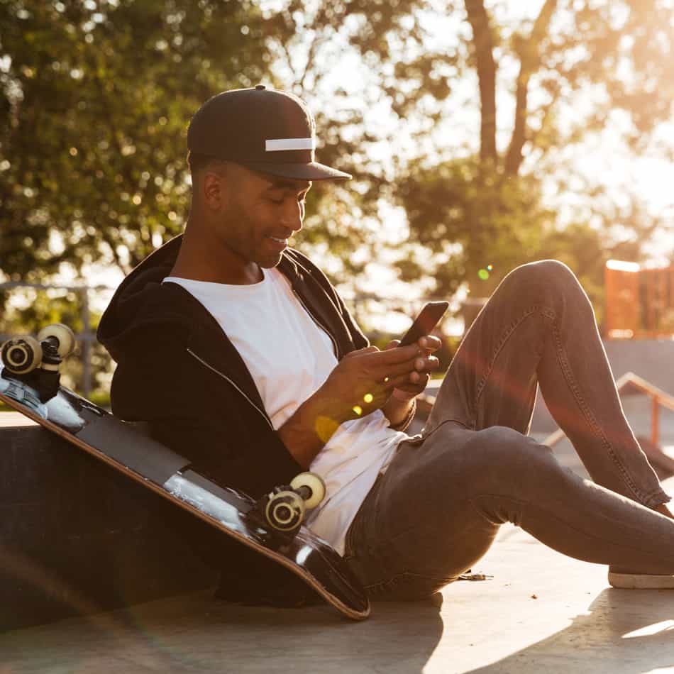 Guy looking at iphone sitting next to skateboard.